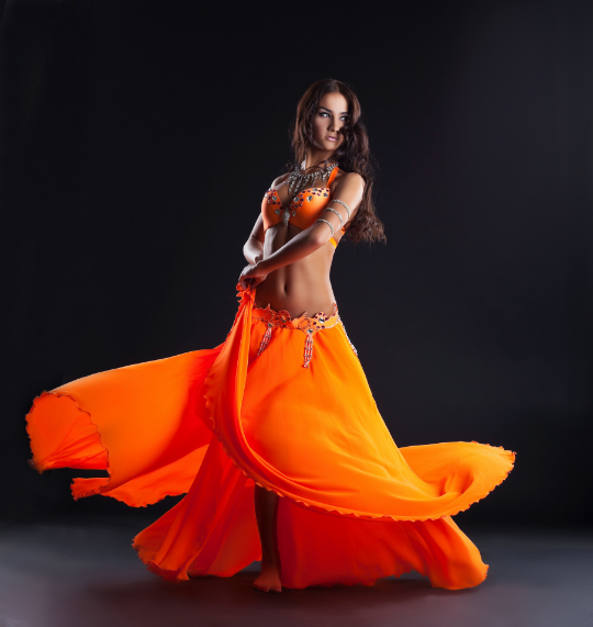 Danseurse | Restaurant Phenicia | Restaurant Phenicia Belly dancer in a bright orange costume spinning a flowing skirt against a dark studio backdrop, arms crossed in front of her torso.